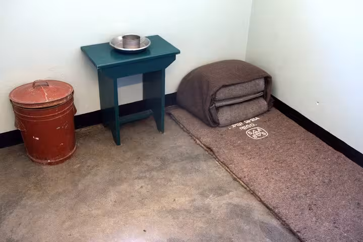 Sparse prison cell interior on Robben Island, showcasing a bedroll, small table, and red bucket.