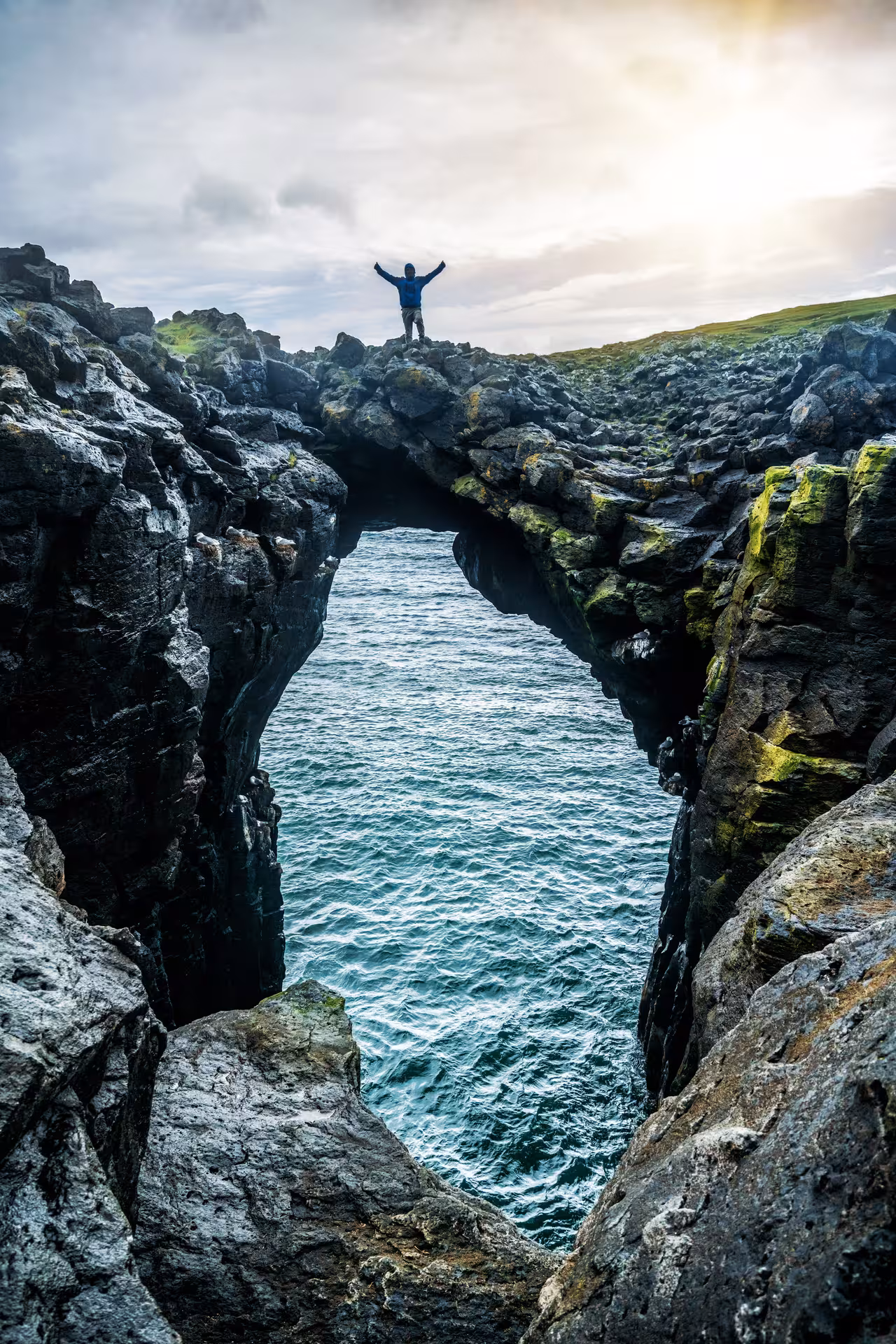 Hiker with arms raised standing on natural rock arch over ocean on Snaefellsnes Peninsula Iceland