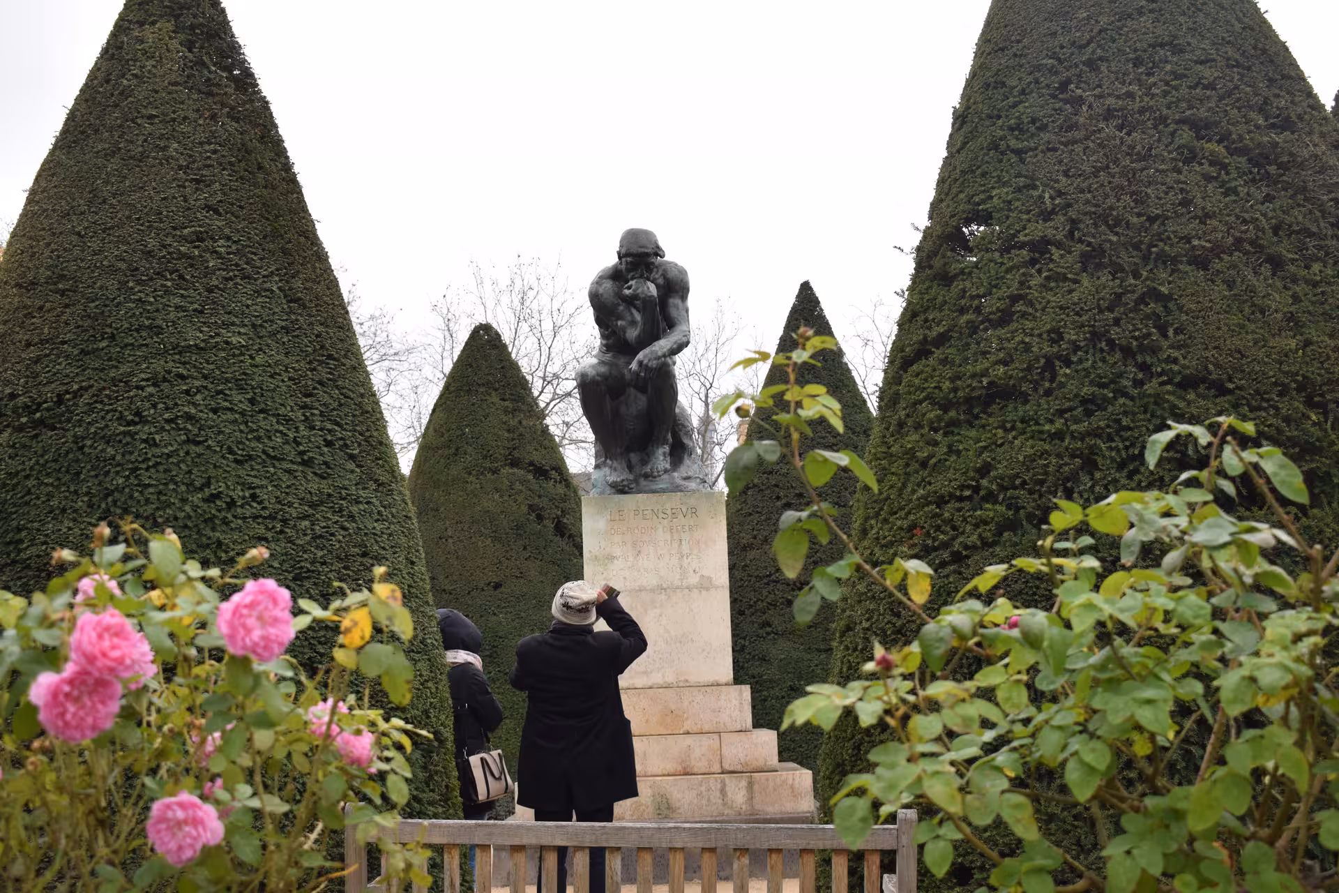 Visitors photographing The Thinker bronze sculpture by Rodin in a garden with trimmed topiary trees and pink roses