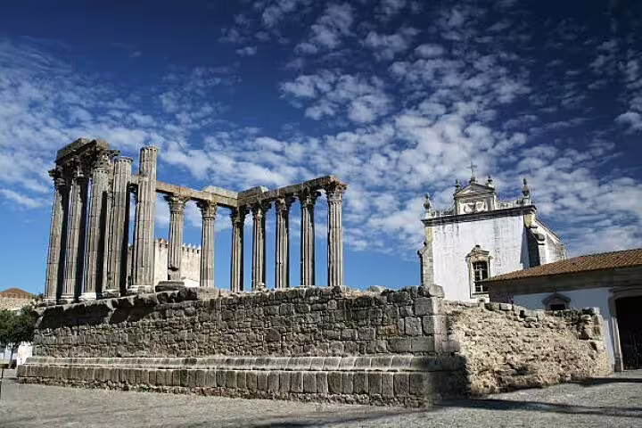 The Roman Temple of Évora, an ancient UNESCO World Heritage site, stands beside a historic church under a cloudy sky.