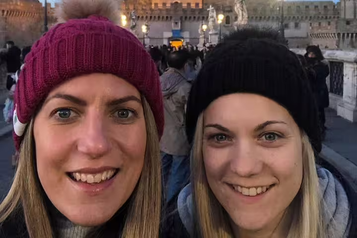 Two smiling women in winter hats on a Rome bridge with Castel Sant'Angelo in background