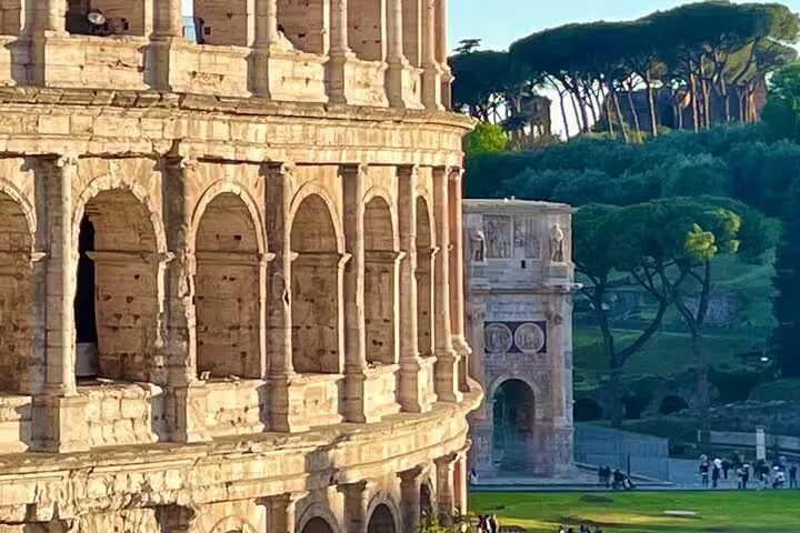 Side view of the Rome Colosseum at sunset with Arch of Constantine and Palatine Hill pines on a panoramic walking tour