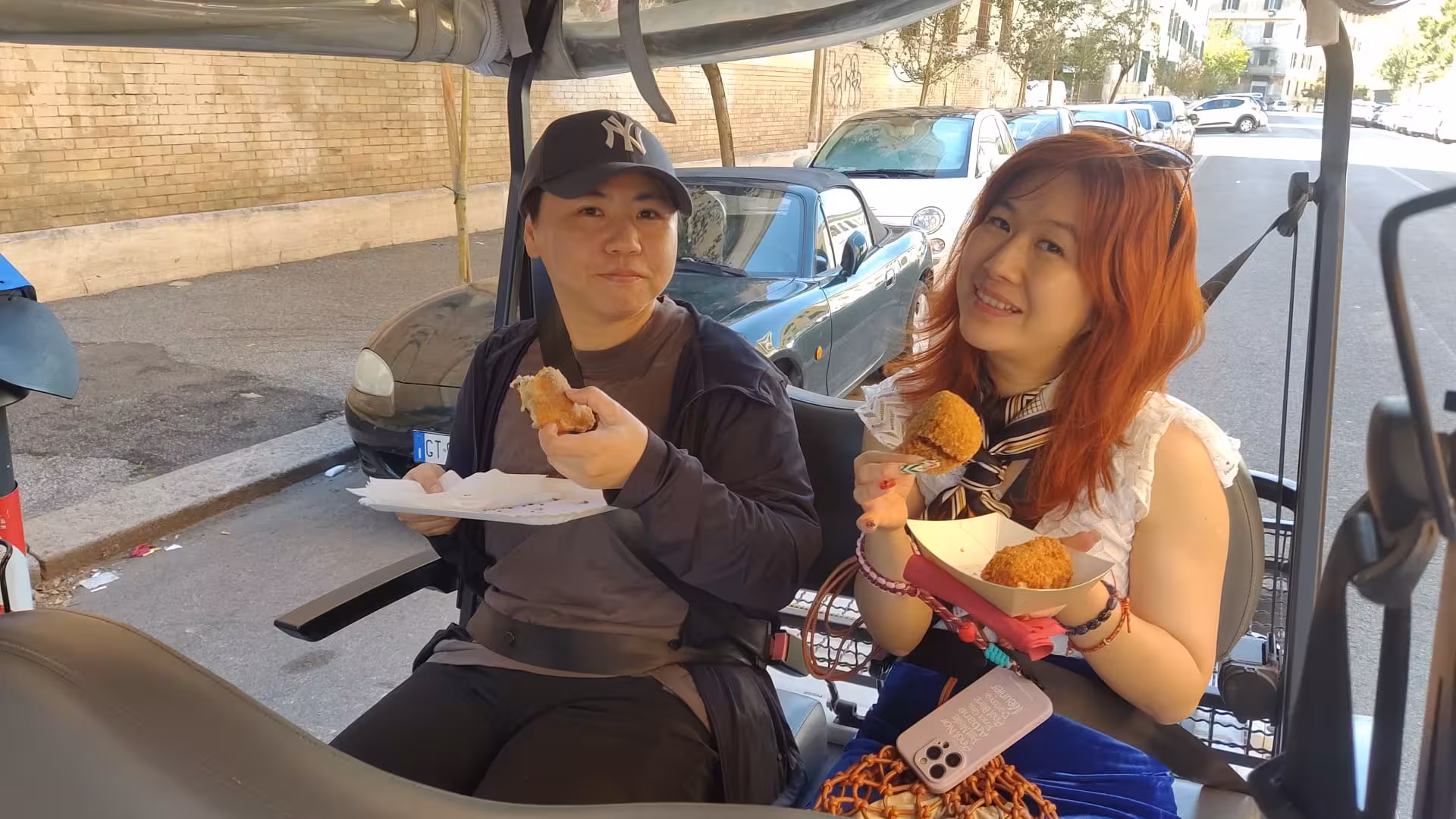 Two tourists enjoy local snacks on a golf cart tour in Rome, perfect for tasting authentic Italian street food.
