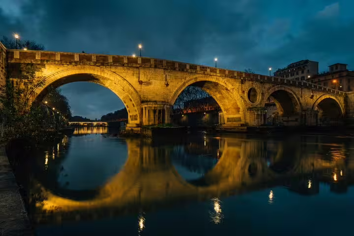 Ancient Roman stone bridge illuminated at night reflecting golden light on the Tiber River in Rome