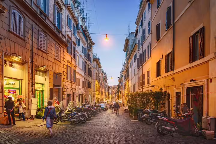 Narrow cobblestone street in Rome at dusk with parked scooters and glowing street lamps