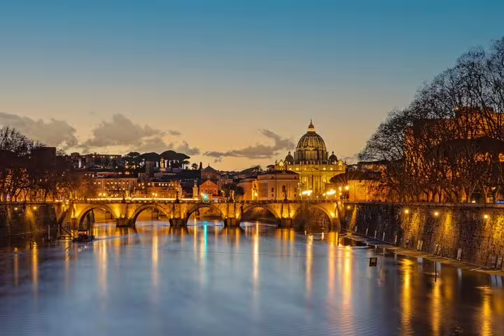 Rome cityscape at dusk with illuminated Ponte Sant'Angelo bridge reflecting on the Tiber River and St. Peter's Basilica