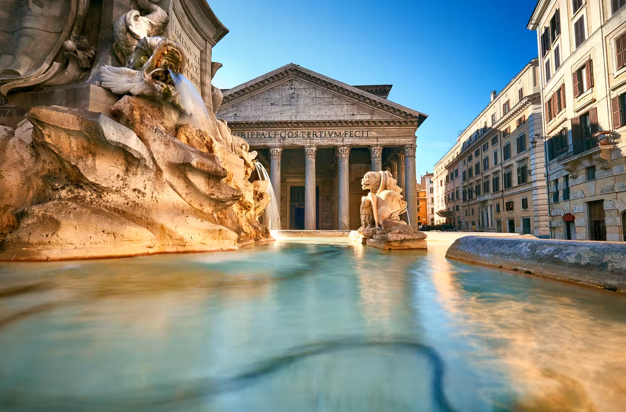 View of the Pantheon in Rome with the Fontana del Pantheon in the foreground under a clear blue sky.