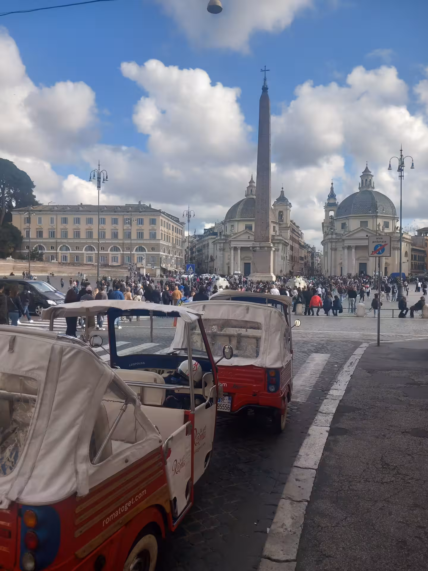 Vibrant tuk-tuks line Piazza del Popolo under a bright sky, capturing the lively atmosphere of Rome's iconic square.