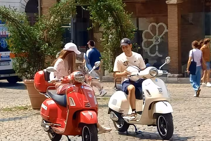 Two friends on red and white Vespas in a sunny Roman plaza, highlighting the fun of a guided Vespa tour.