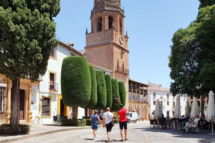 Tourists stroll past the historic clock tower and manicured trees in Ronda's charming plaza during a private walking tour.