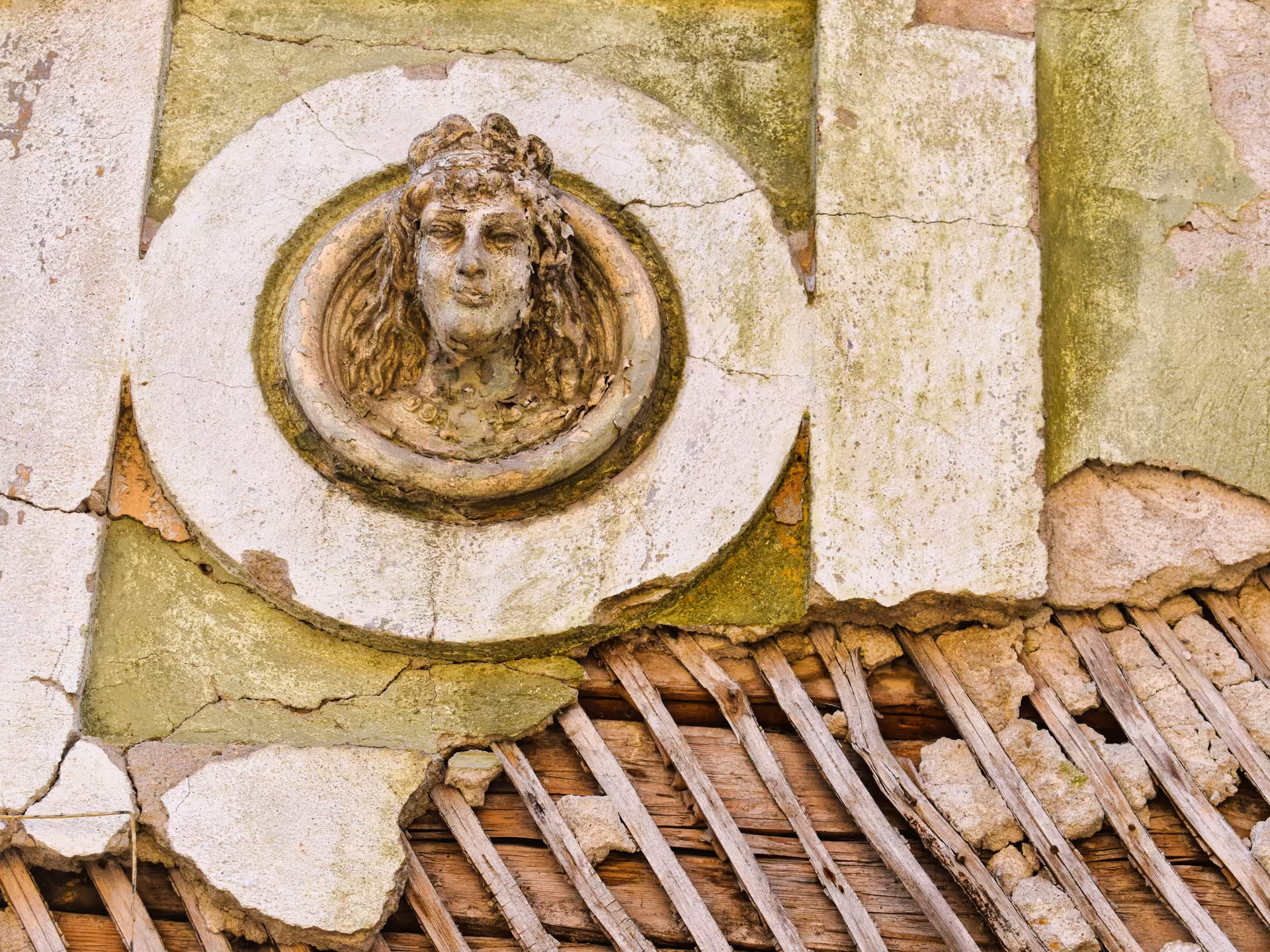 Weathered stone relief of a female face on crumbling plaster wall with exposed wooden laths, Rosia Montana