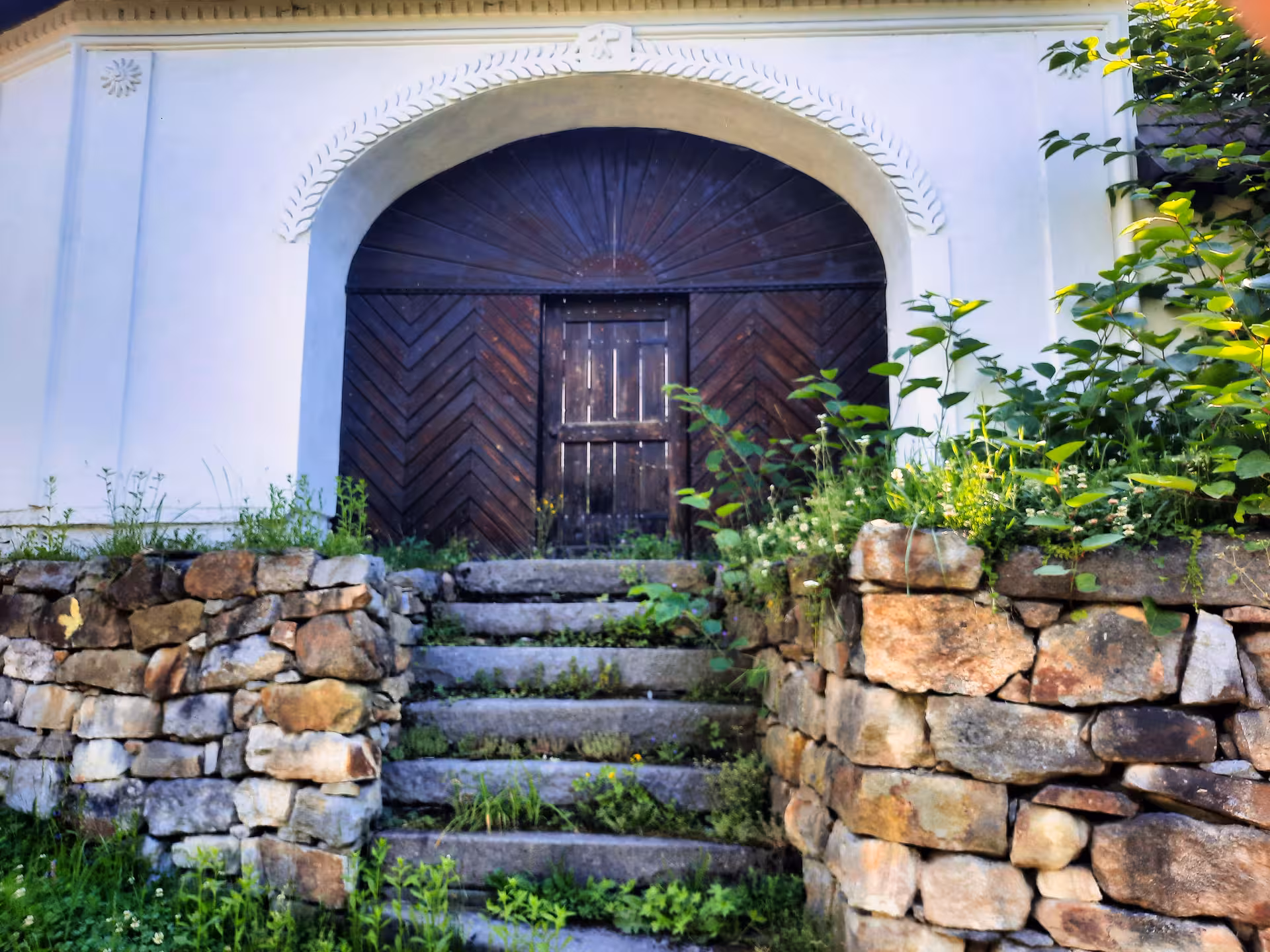 Ornate arched wooden door with chevron pattern at historic Rosia Montana building, flanked by stone walls and steps
