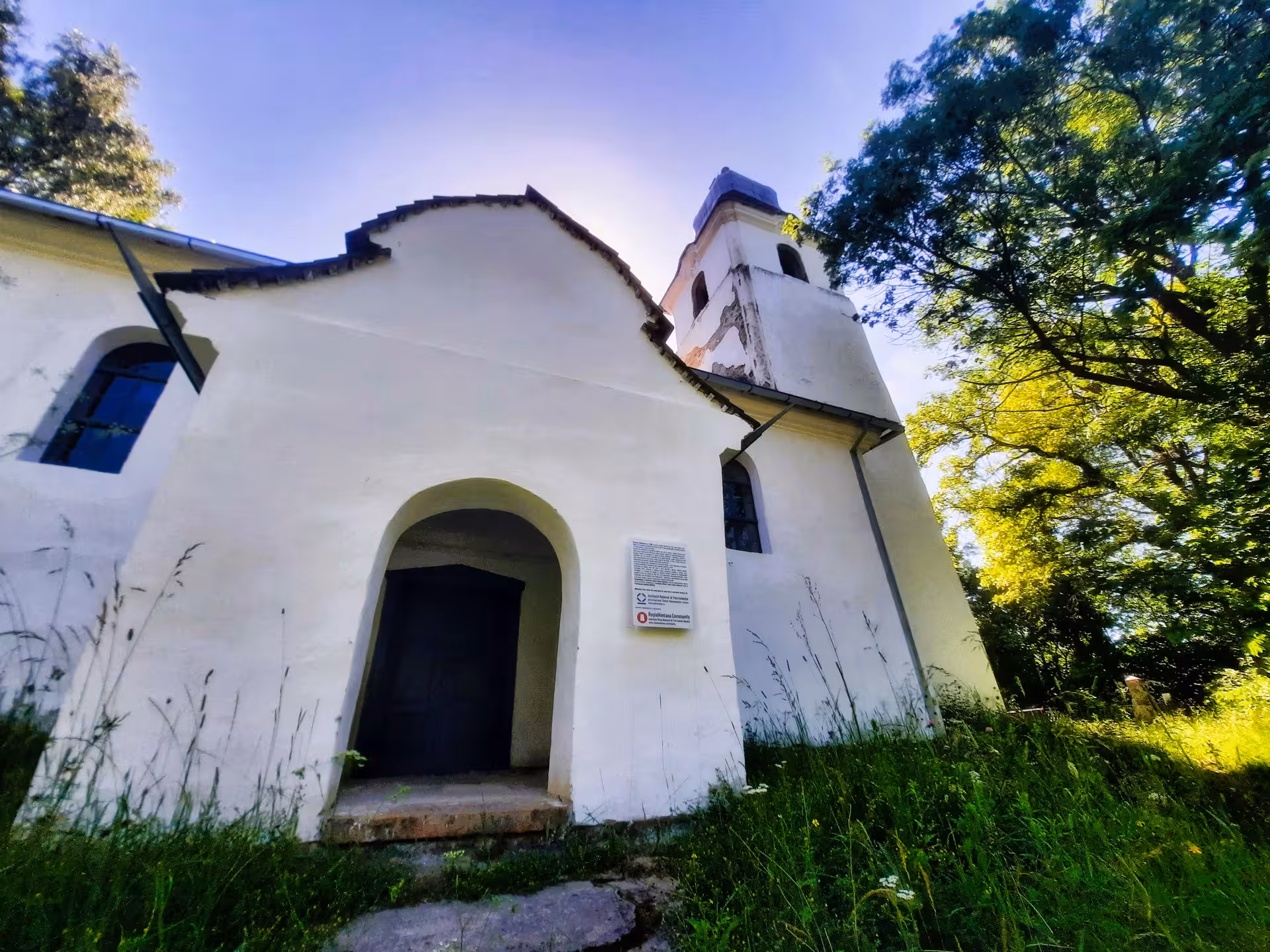 White historic church with arched entrance and bell tower surrounded by lush greenery in Rosia Montana