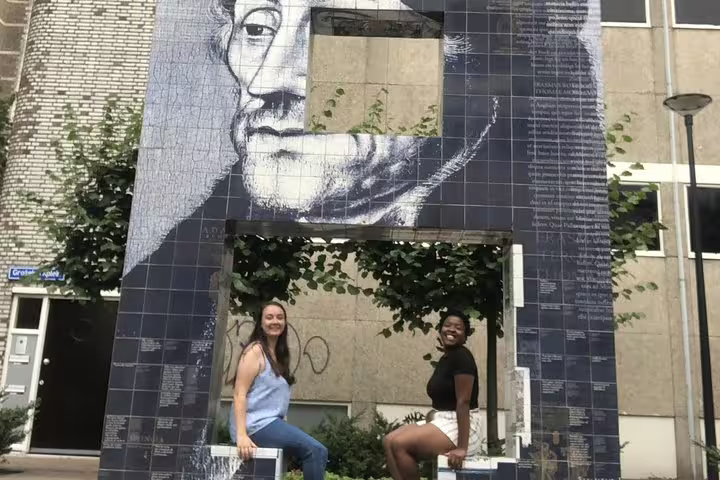 Two women posing in front of a large tiled mural portrait on a Rotterdam city street