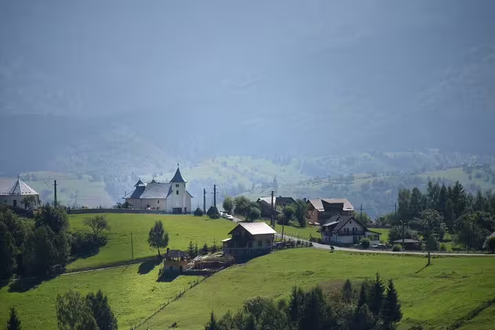 Green hillside Romanian mountain village with white church and traditional houses in misty Carpathian landscape