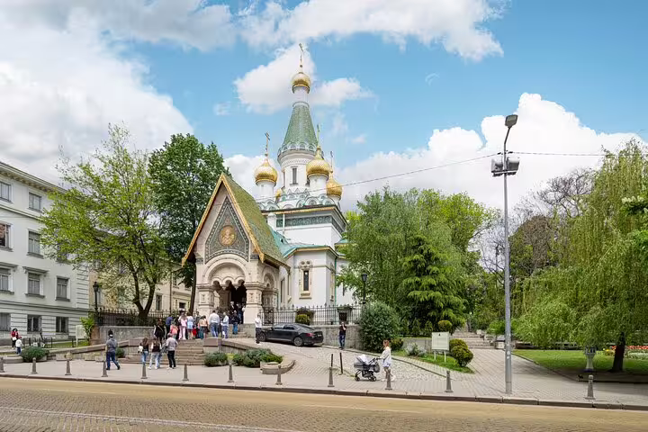 Russian Church with golden domes in Sofia, Bulgaria, surrounded by trees and visitors on a sunny day