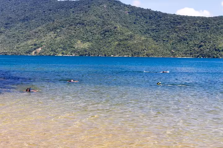 Visitors snorkeling in the clear, turquoise waters of Saco Mamanguá Fjord with lush green hills in the background.