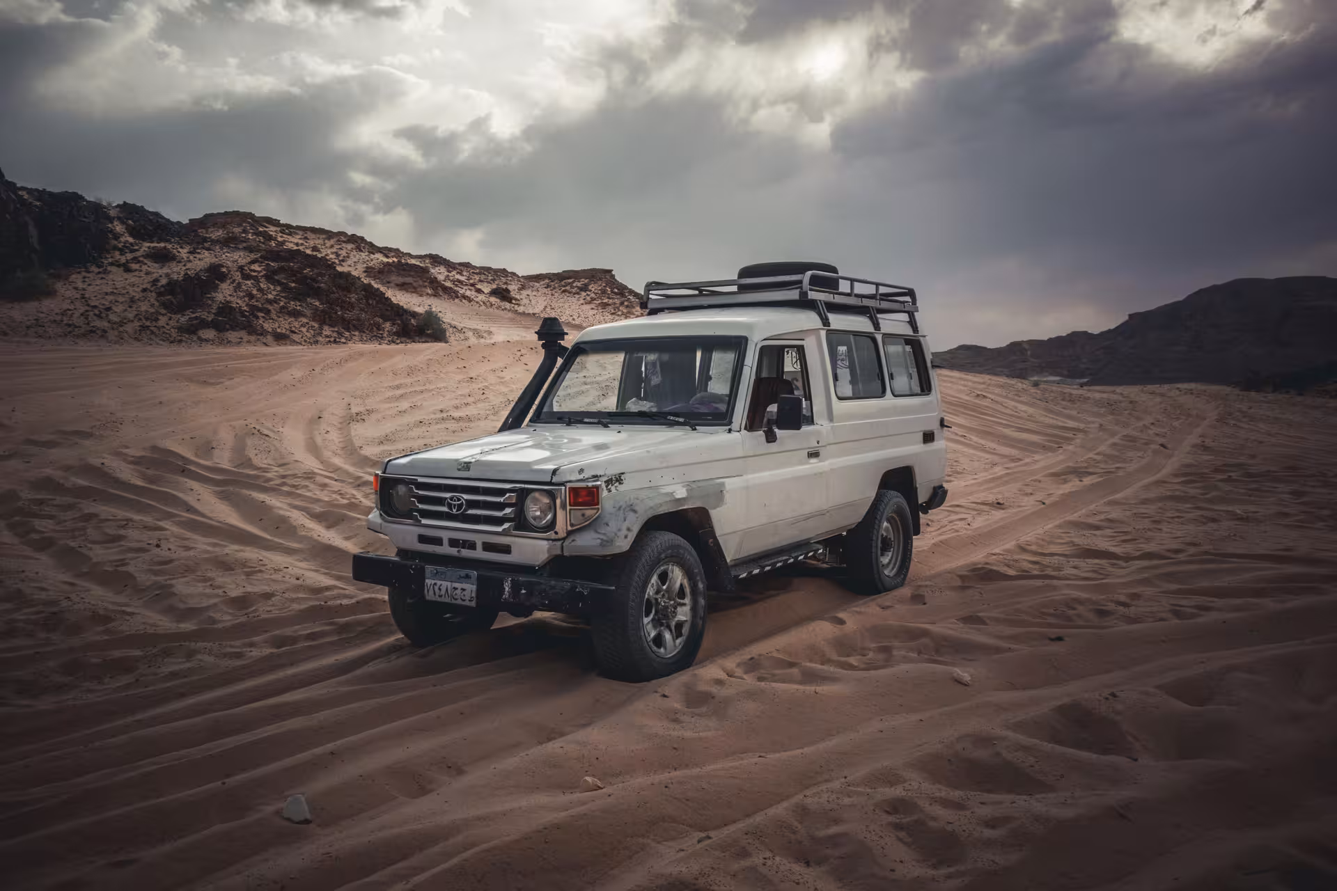 White Toyota Land Cruiser 4x4 jeep driving through red sand desert dunes under cloudy sky