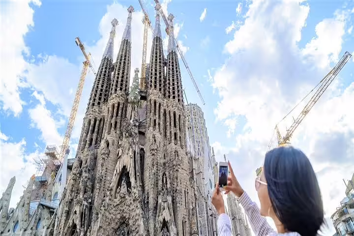 Tourist photographing Sagrada Familia's iconic spires under blue skies, highlighting Barcelona's architectural marvel.