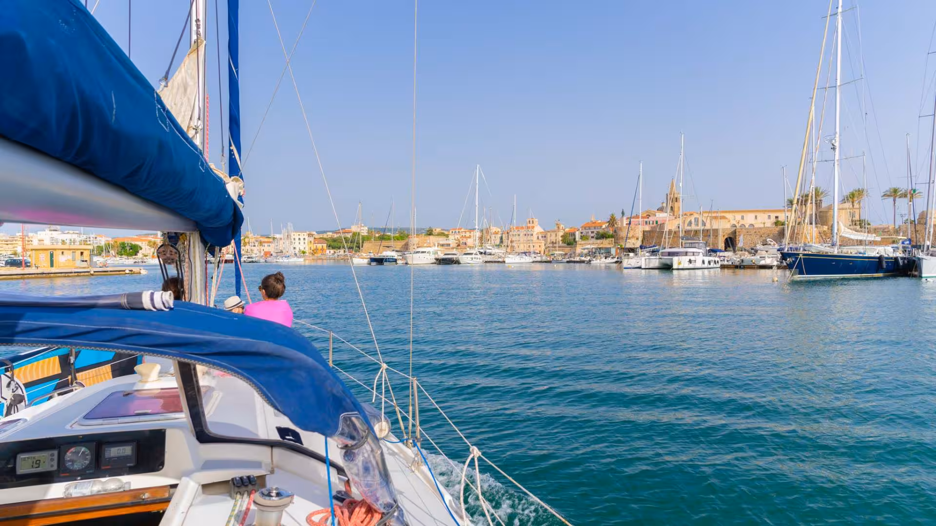 Sailboat approaching Alghero harbor with scenic views of Porto Conte Park and marina.