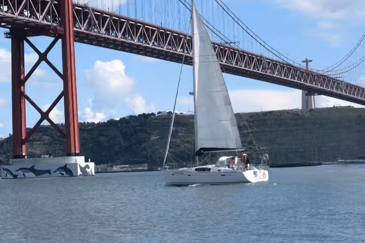 Sailboat gliding under Lisbon's iconic red bridge on a sunny morning Tagus River tour, showcasing Portugal's vibrant scenery.