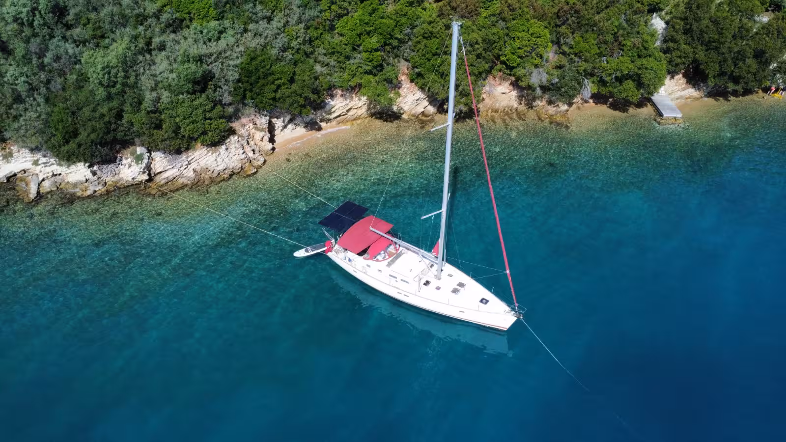 White sailboat anchored in crystal-clear turquoise waters near a rocky tree-lined coastline, viewed from above.