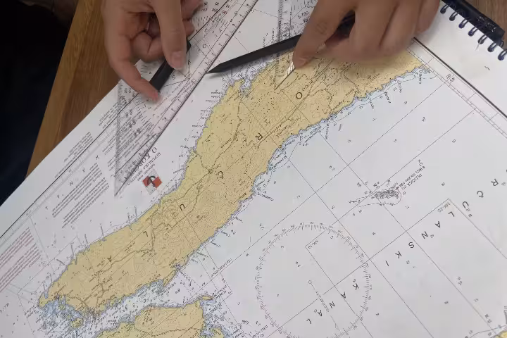 Hands plotting a route on a nautical chart with ruler and pencil, part of an online 9-hour sailing basics class