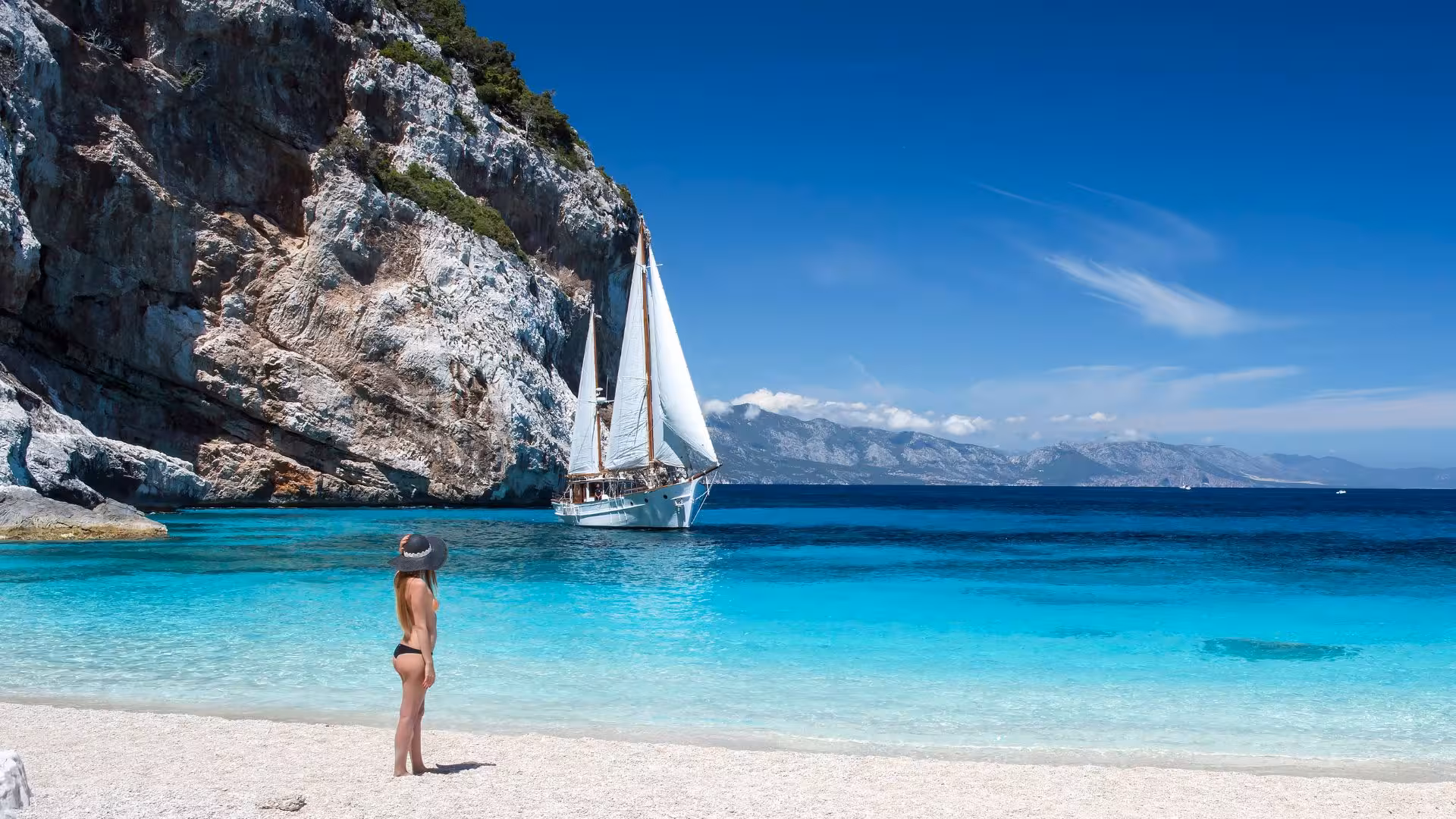 Woman on a sandy beach watches a sailboat with white sails near the dramatic cliffs of the Gulf of Orosei in Cala Gonone.