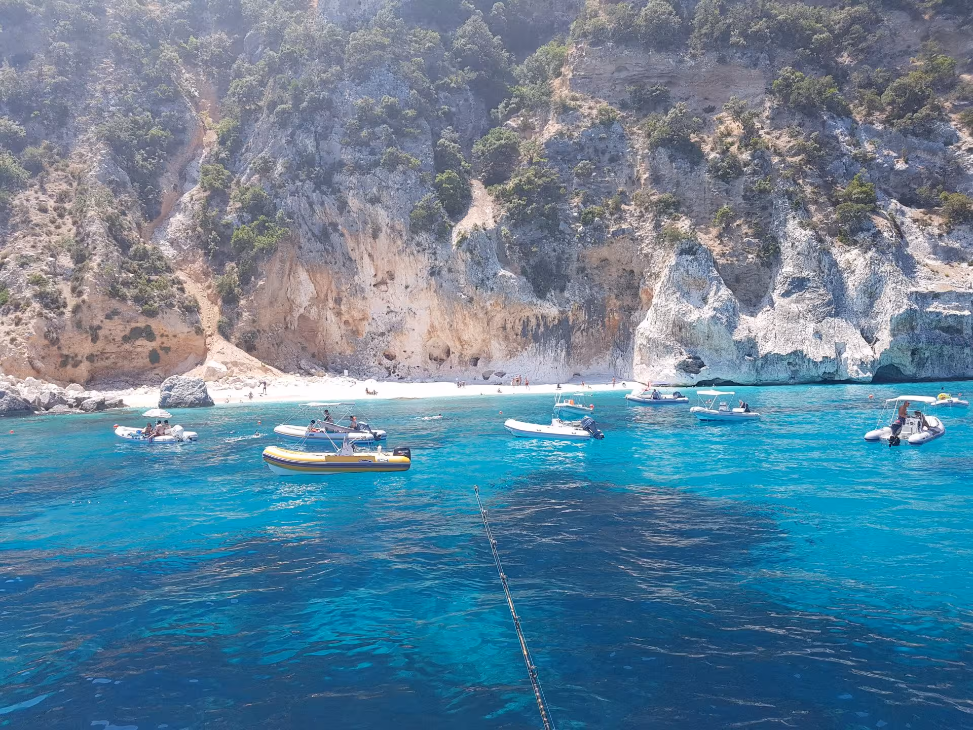 Multiple boats anchored in the vibrant blue waters near a rocky coastline in the Gulf of Orosei, Sardinia.