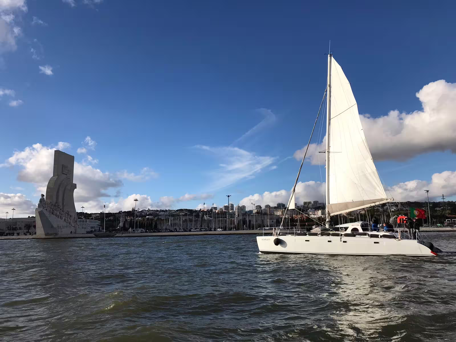 Sailing catamaran Scarlett cruising near Lisbon's iconic Discoveries Monument under a clear blue sky.