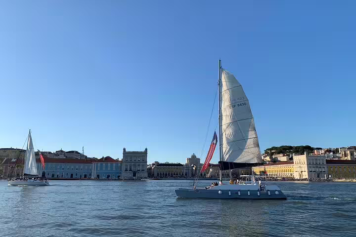 Sailing catamaran Vertigo gliding on Lisbon's Tagus River with iconic cityscape in the background on a sunny day.