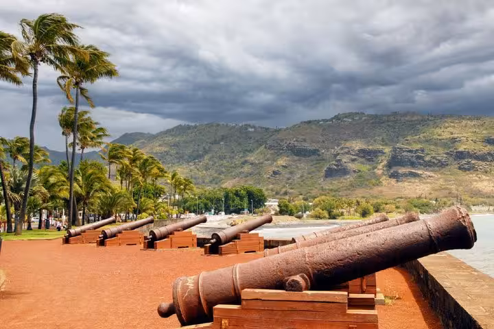 Historic cannons lined up along Saint-Denis waterfront with palm trees and green mountains under stormy sky