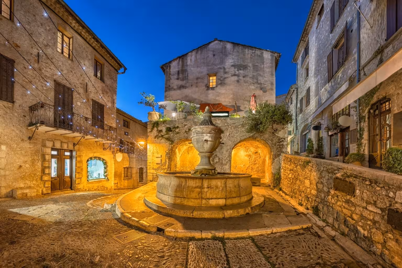 Illuminated stone fountain in the medieval village square of Saint-Paul-de-Vence at dusk, Provence, France