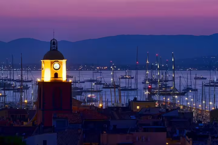 Illuminated clock tower of Saint-Tropez at dusk with marina and sailboats in background