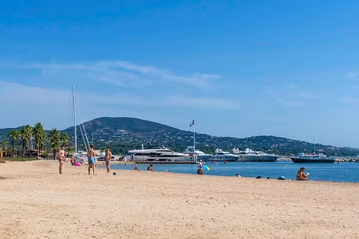 Sandy beach with yachts moored at Saint-Tropez harbor, palm trees and green hills in background