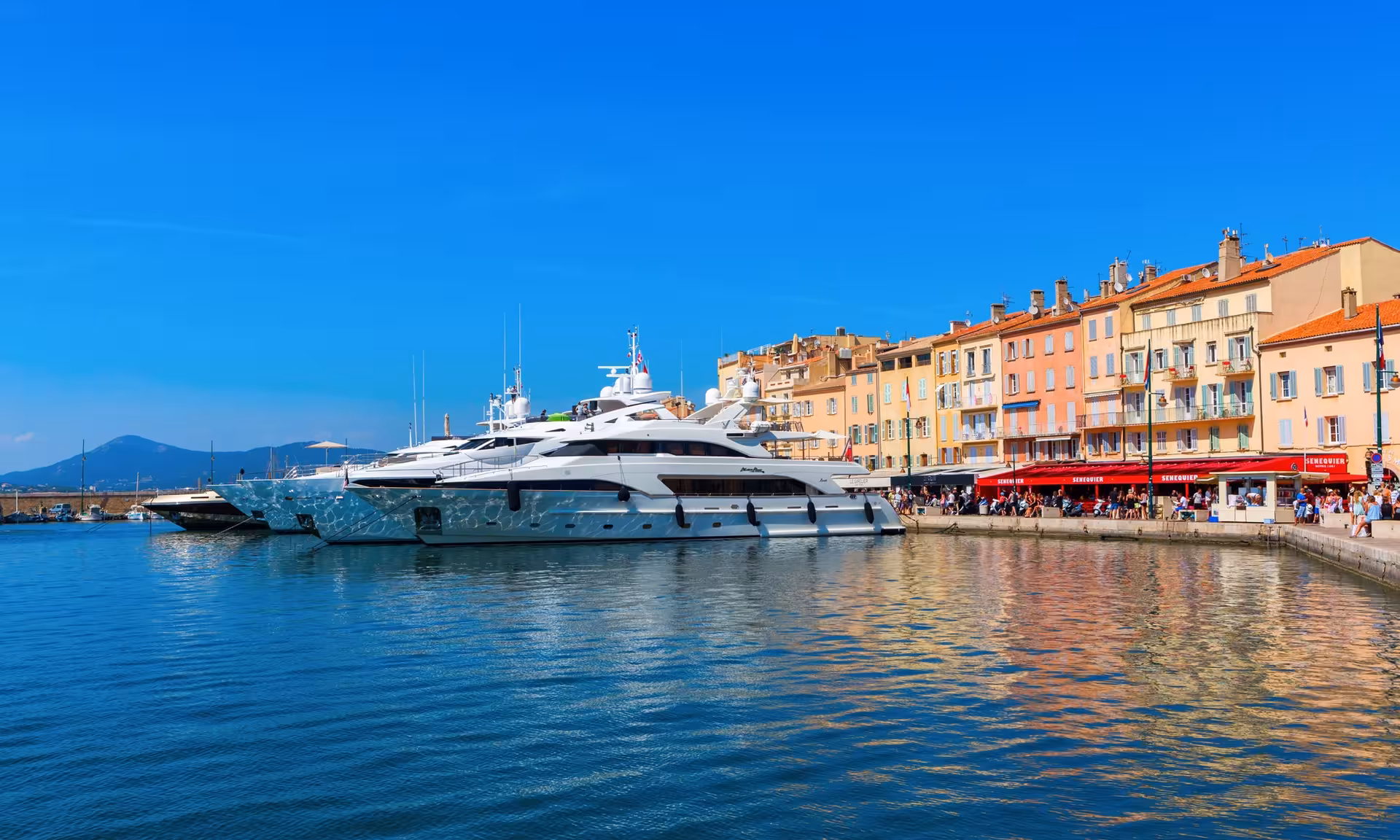 Luxury yachts moored in Saint-Tropez harbor with colorful waterfront buildings and Senequier café on sunny day