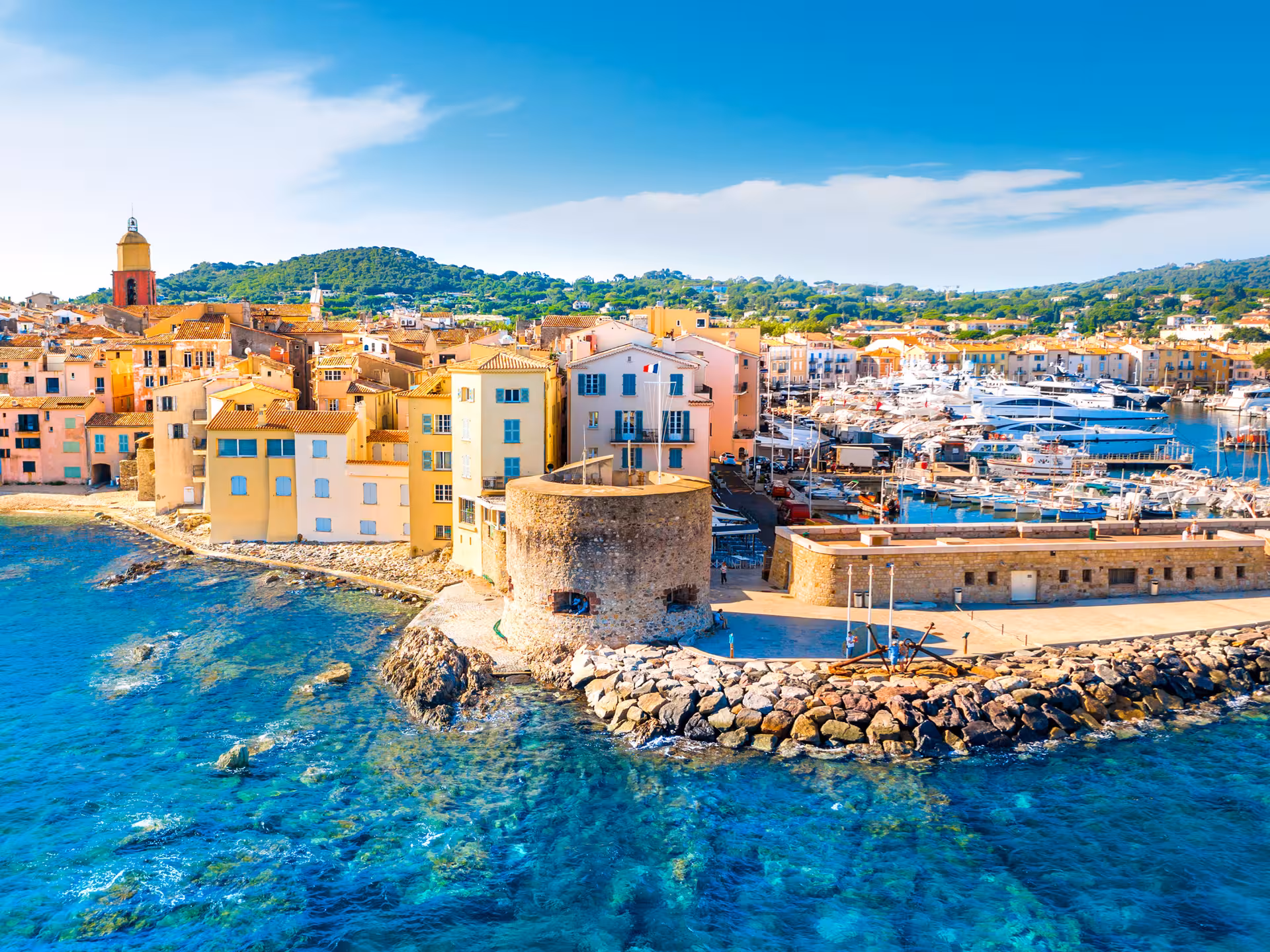 Aerial view of Saint-Tropez harbor with colorful buildings, ancient stone tower, and luxury yachts on turquoise water