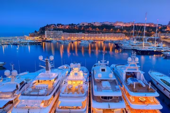 Luxury yachts docked at a glowing marina at dusk with hillside buildings in the background