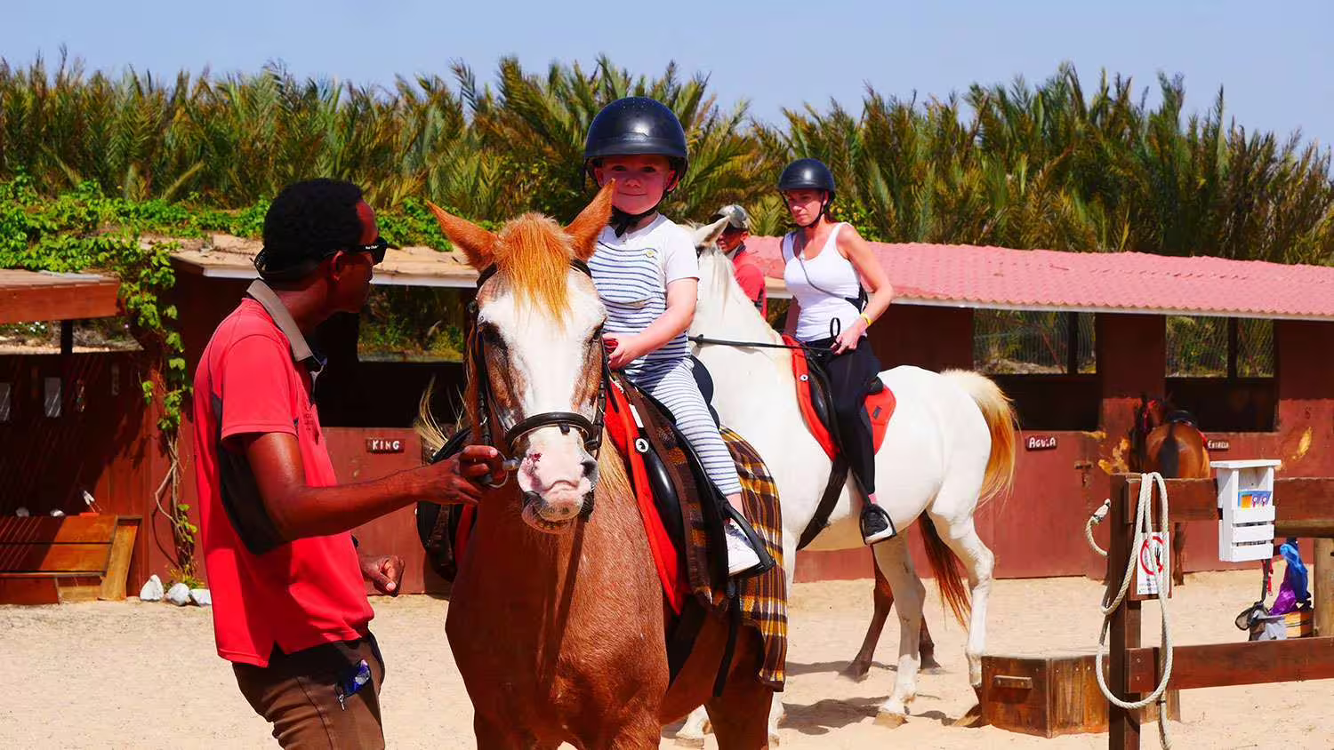 Child and adult horseback riding in a sandy arena with a guide, set against lush green palm trees on Sal Island.