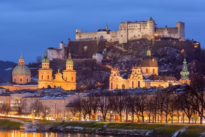 Salzburg old town skyline at dusk with Hohensalzburg Fortress illuminated above historic churches and the Salzach River