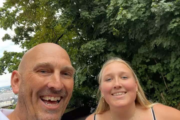 Smiling man and young woman taking a selfie outdoors in Salzburg Old Town with green trees behind them