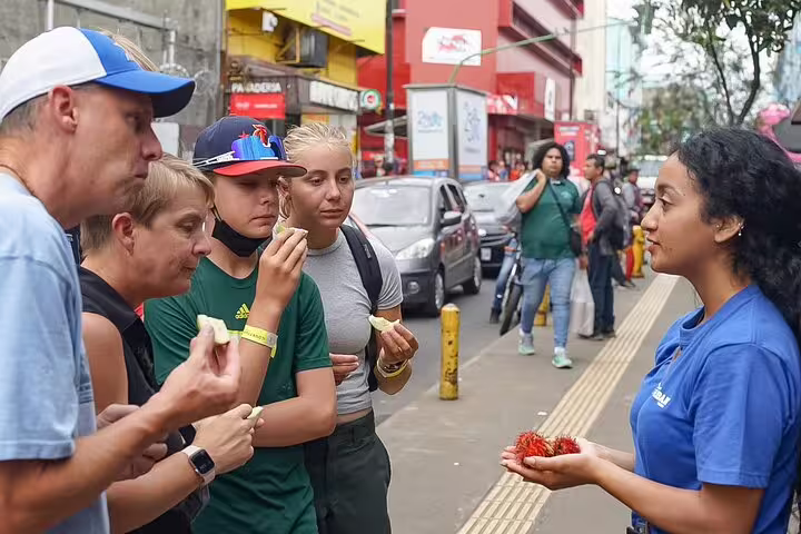 Tourists enjoy exotic fruits with a guide on a bustling San Jose street during a food tour.