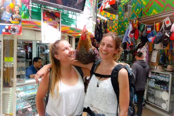 Two women smiling with a rooster in a vibrant San Jose market during the Bites and Sights Walking Food Tour.