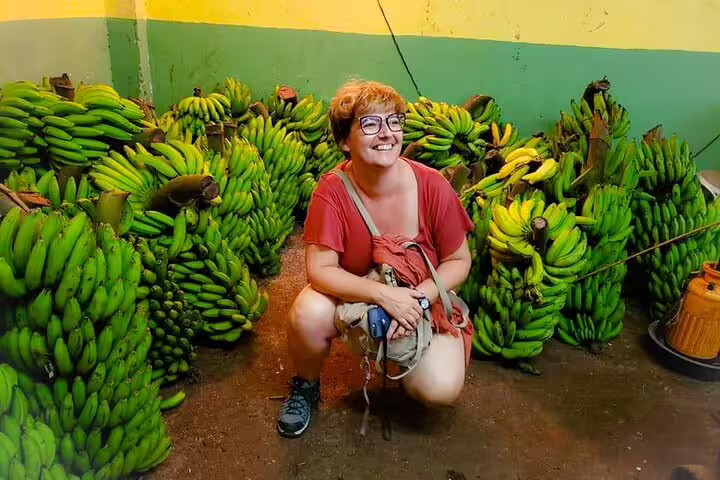 A visitor poses happily amidst piles of fresh bananas at a bustling San Jose market during the walking food tour.