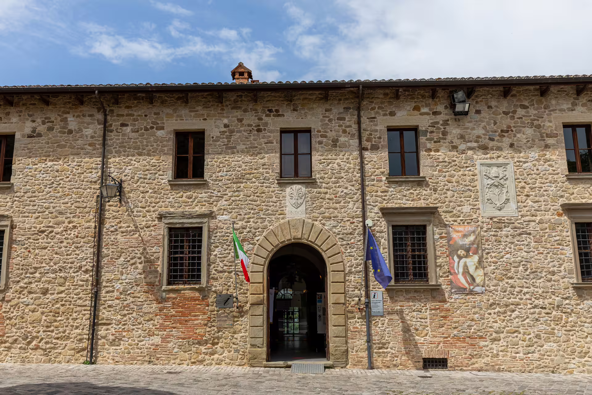 Front view of San Leo fortress entrance with Italian and EU flags, showcasing historical architecture.