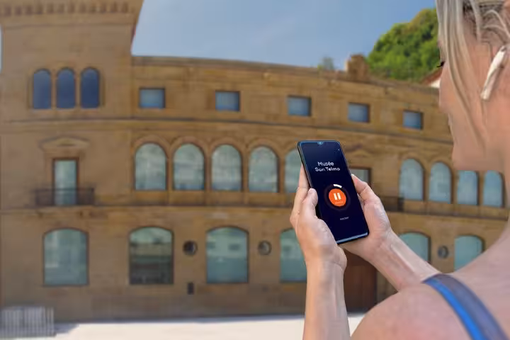 Tourist holding smartphone with San Telmo Museum audio tour app paused in front of historic building in San Sebastian