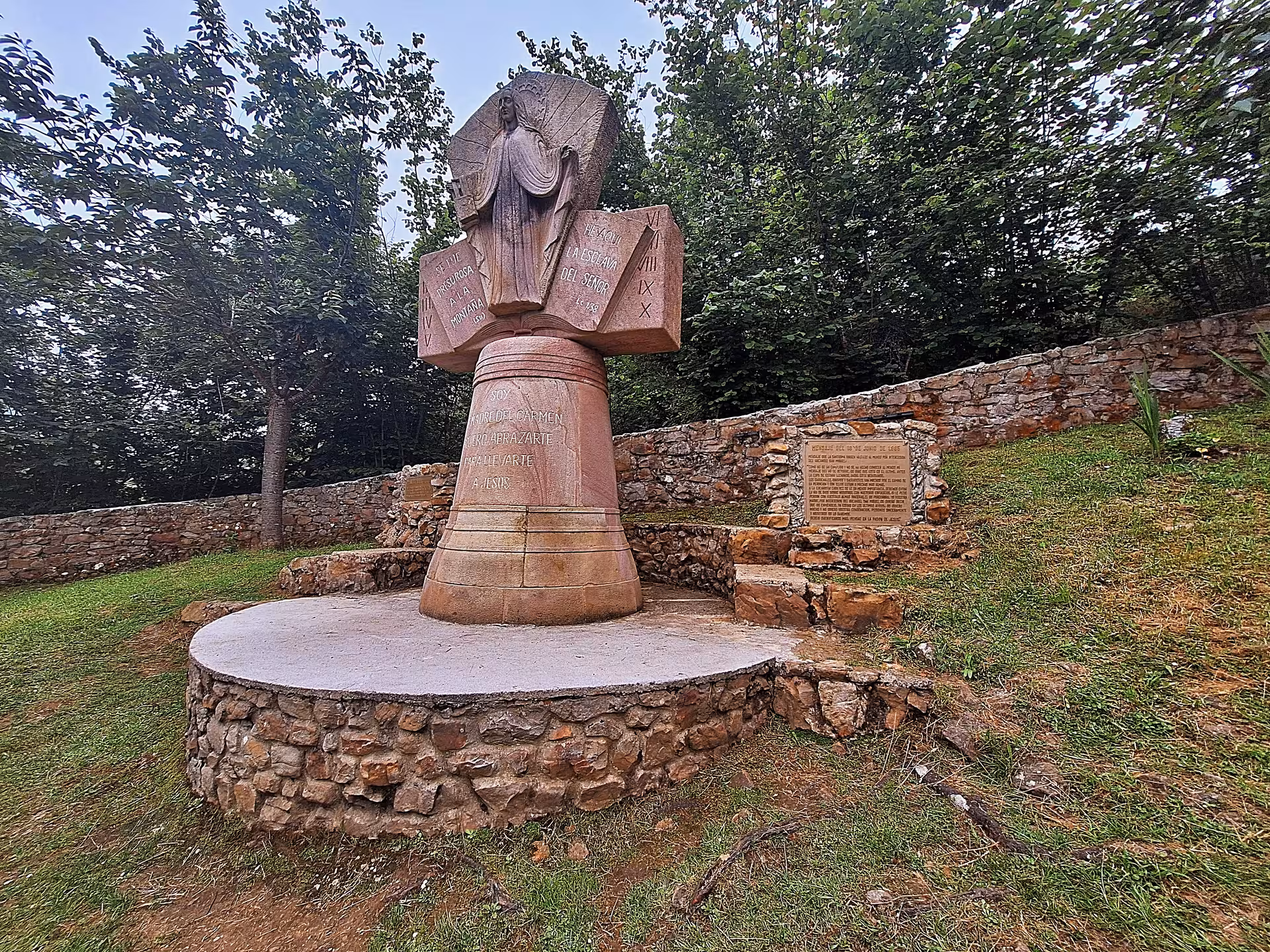 Visit the serene monument in San Sebastian De Garabandal surrounded by lush greenery and historical inscriptions.