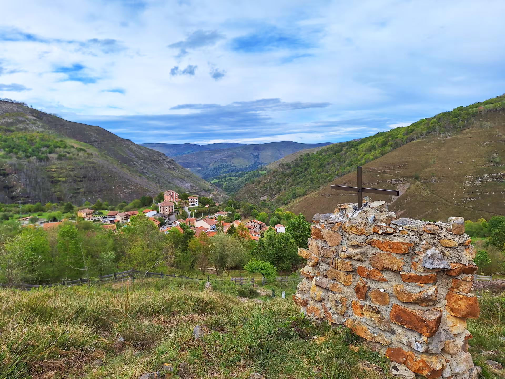 Scenic view of San Sebastian De Garabandal village nestled in lush hills with a stone cross in the foreground.