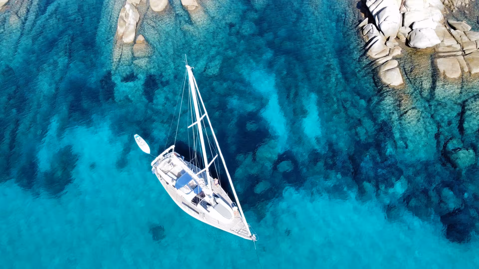 Aerial view of a sailboat in crystal-clear turquoise waters near rocky shores on the San Teodoro sailing tour.