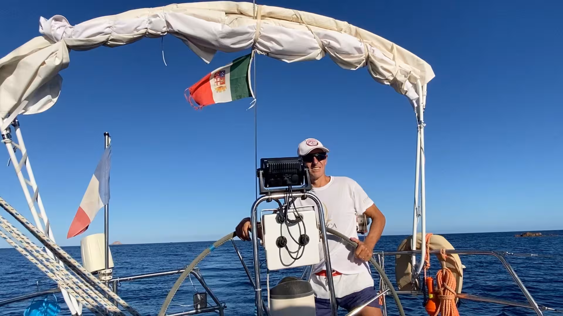 Captain steering sailboat under clear skies on San Teodoro sailing tour with Italian flag fluttering.
